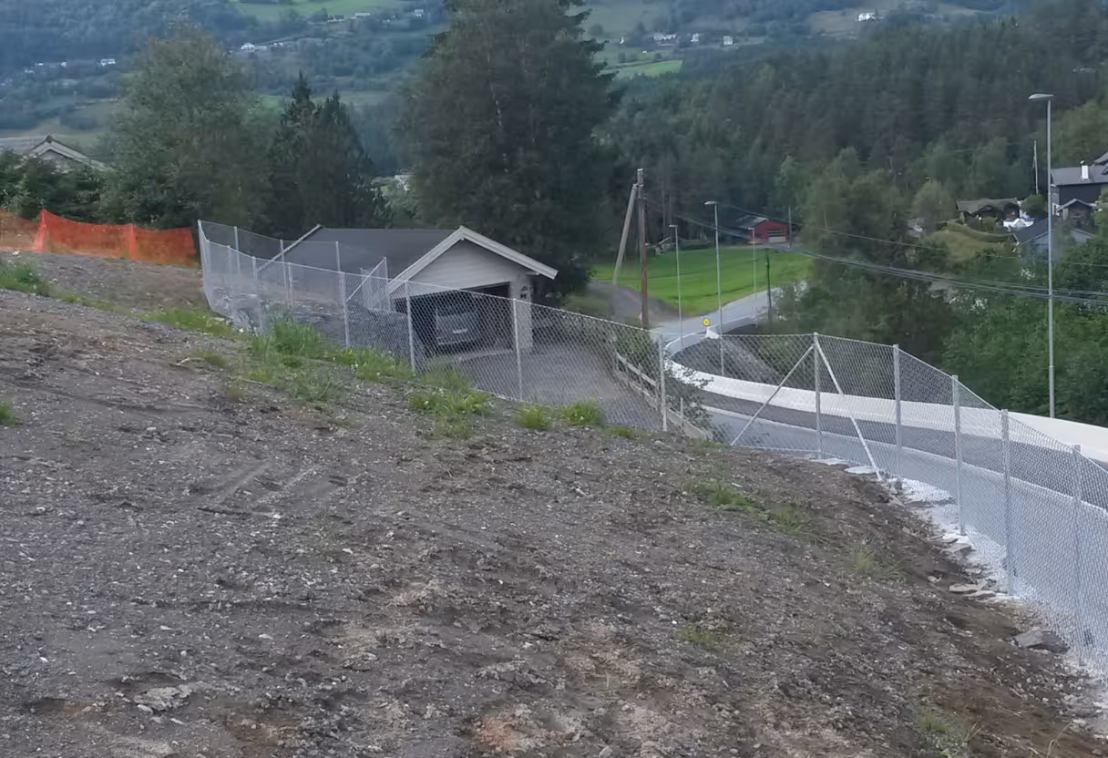 Kjettinglenkegjerde rundt en tomt med skrånende jord og en rekke hus i bakgrunnen.