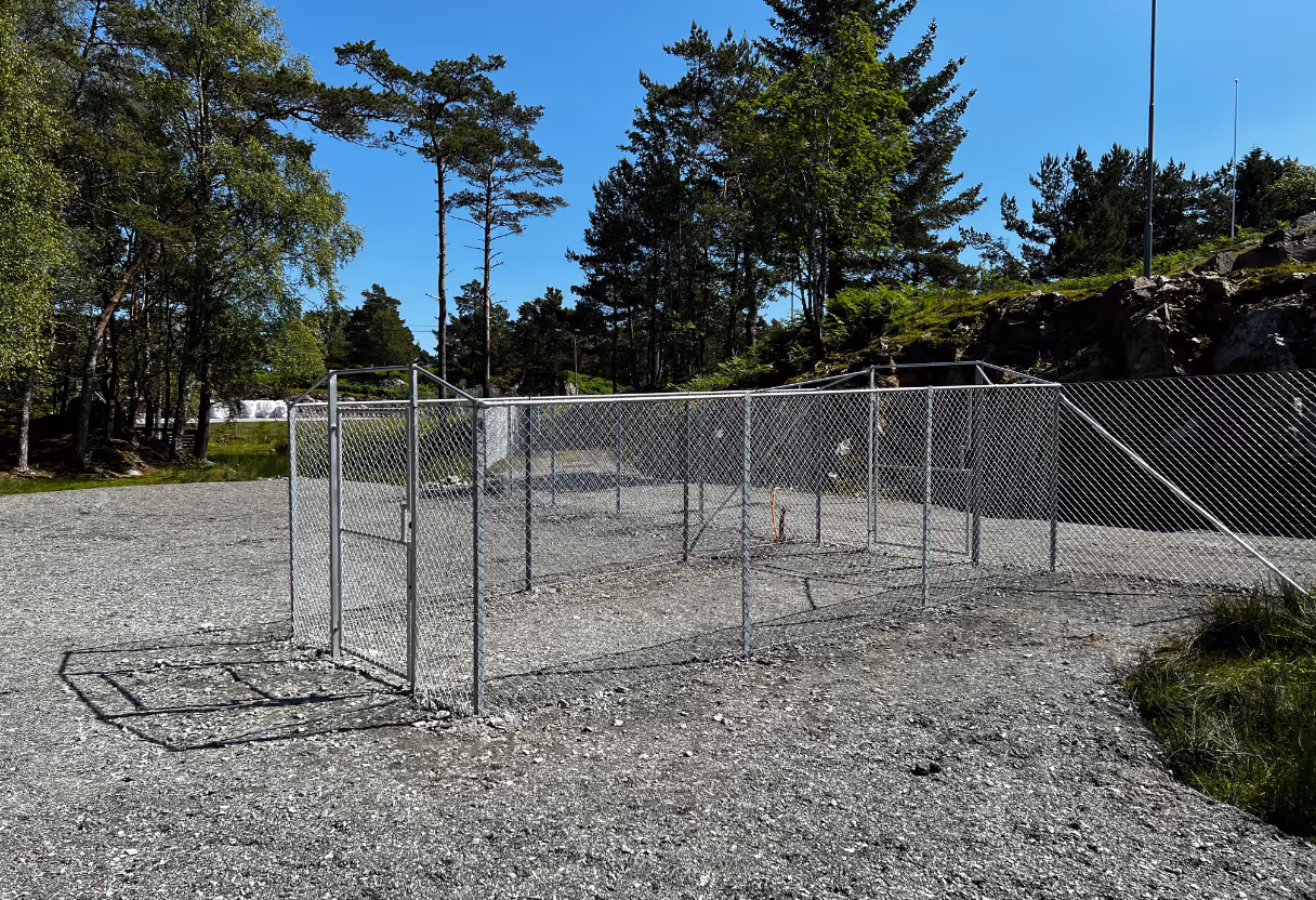 Metallgjerde i netting med port plassert på grusområde omgitt av trær og blå himmel.
