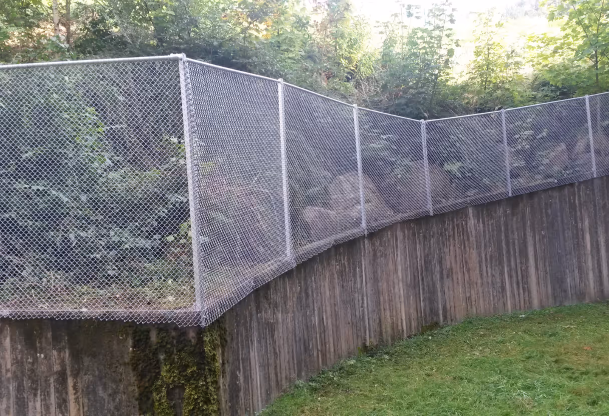 Stål nettinggjerde montert over trevegg langs en gressplen og skogområde bak.