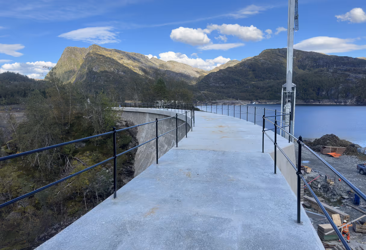 Betongrekkverk langs svingete vei med fjell og vann i bakgrunnen under en blå himmel.