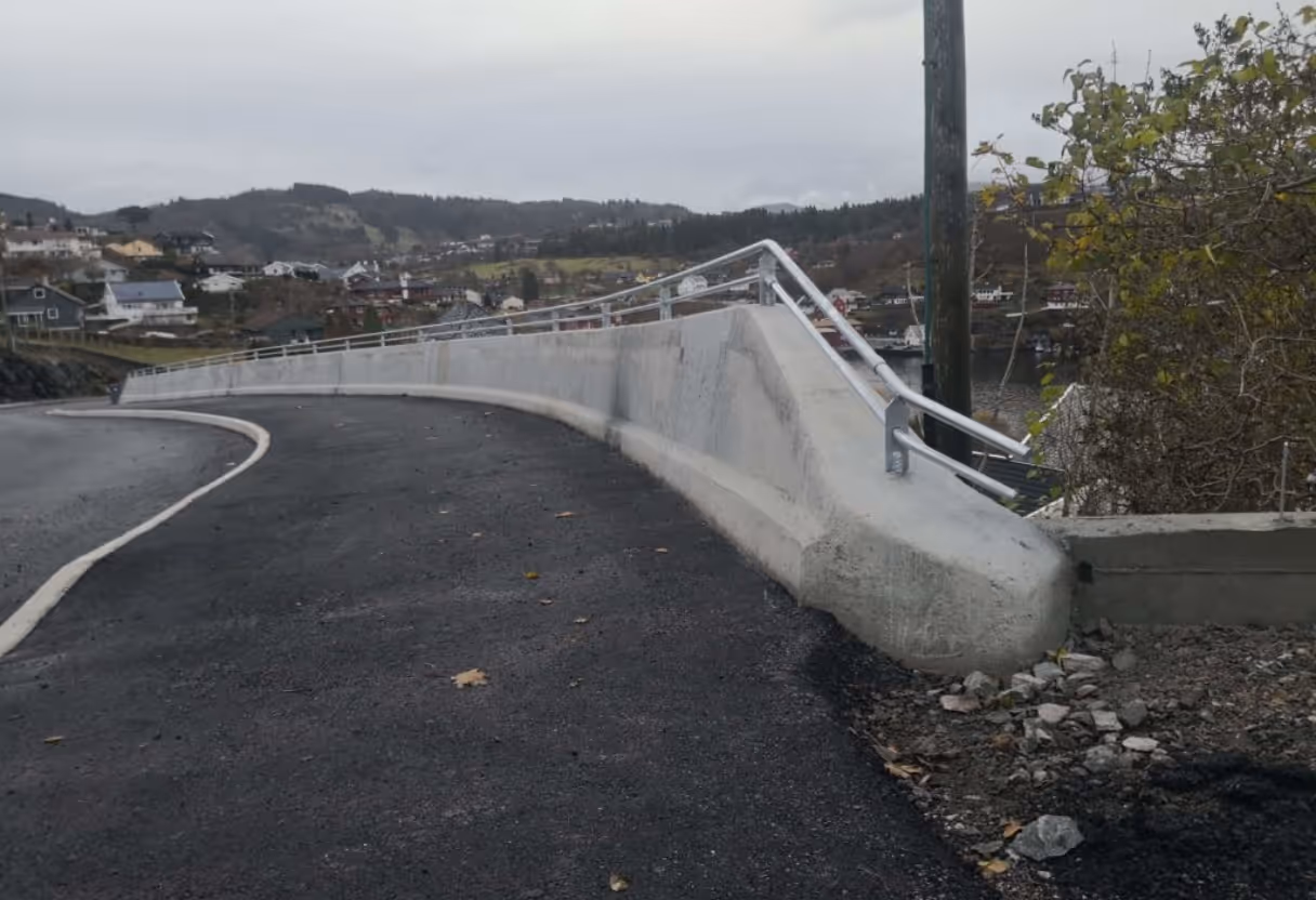 Sykkelvei med mørk asfalt langs en betongbarriere med rekkverk, omgitt av hus og skog.