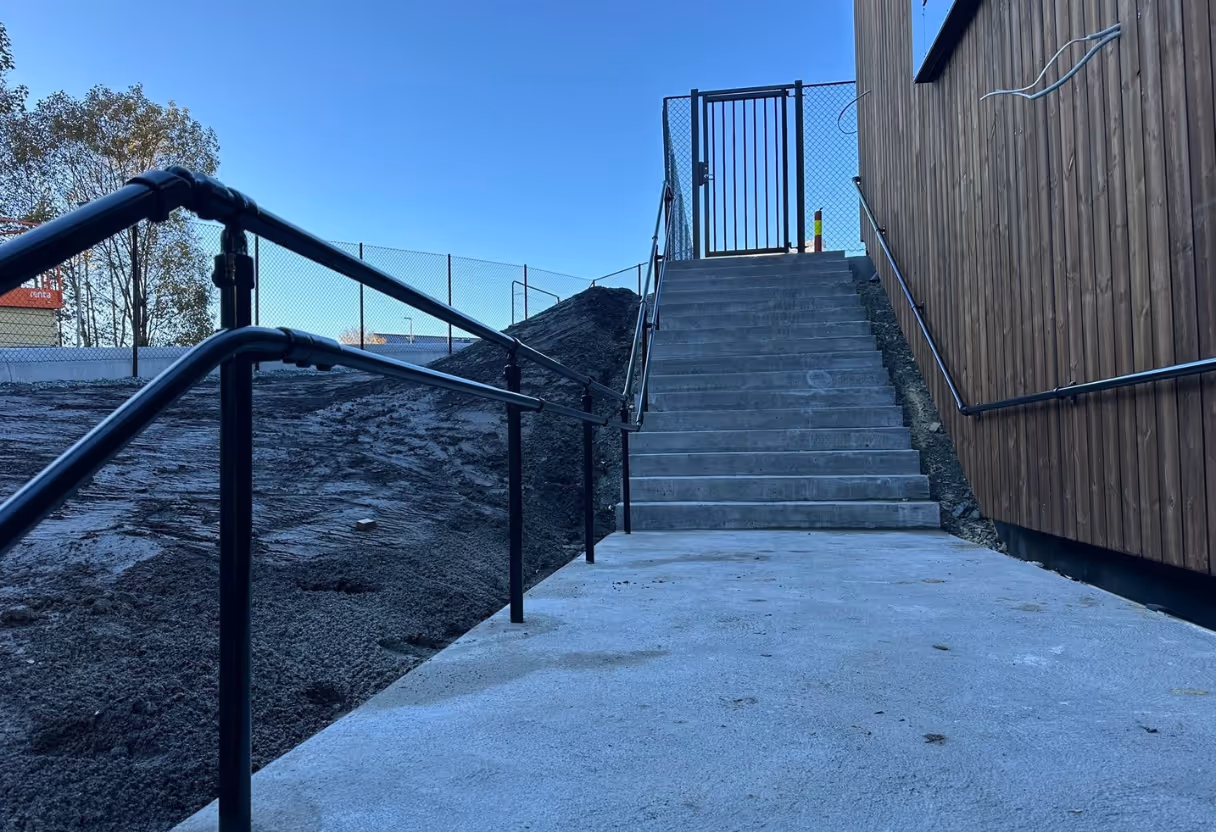 Grå betongtrapp med svarte rekkverk ved siden av et trevegget bygg under klar blå himmel.