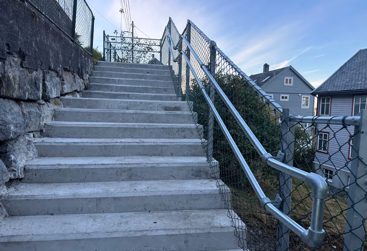 Betongtrapp med solide metallrekkverk og nettinggjerde, med hus i bakgrunnen under blå himmel.