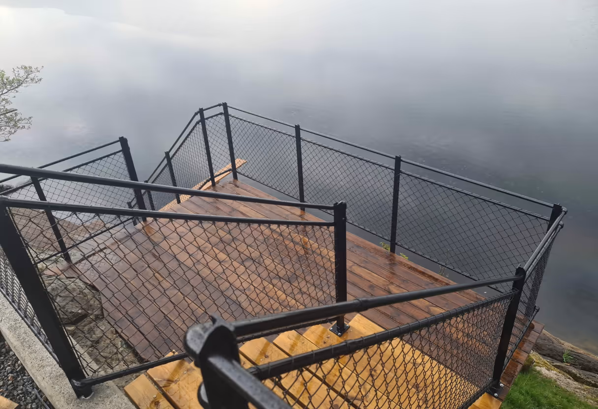 Treppe med svarte rekkverk ned mot vann med speilblank overflate og små steiner på siden.
