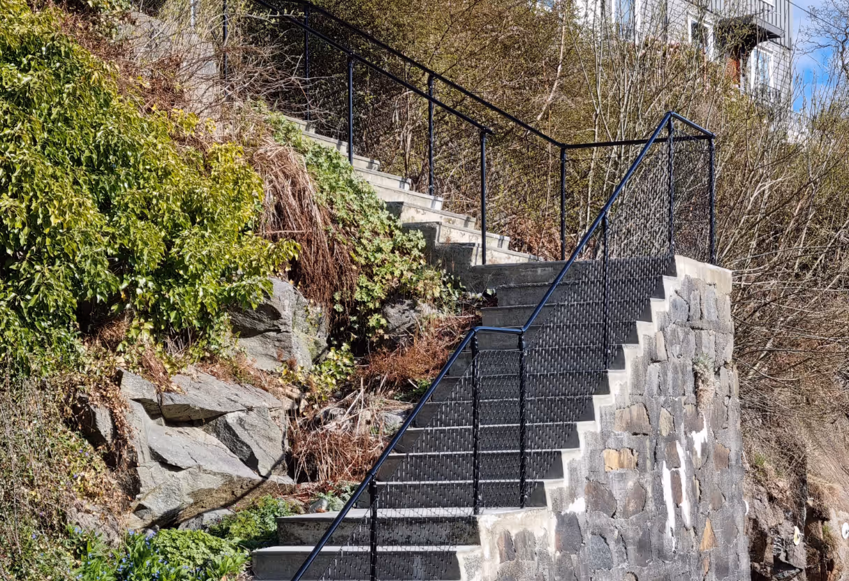 Steintrapper med svart metallrekkverk i naturlig utendørsområde med vegetasjon og bygning i bakgrunnen.