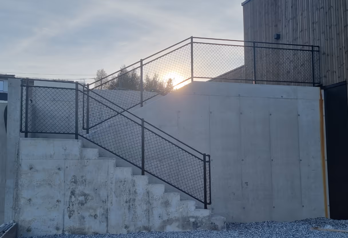 Betongtrapp med svart nettingrekkverk og sol som skinner bak.