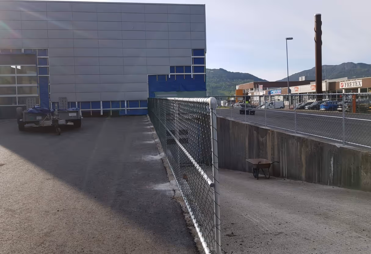Kjedelenke gjerde med hvitt metallramme ved en industriell bygning og en tralle.