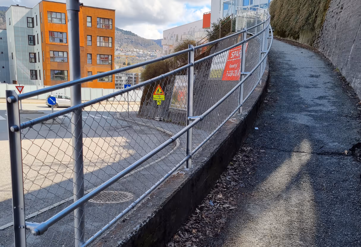 Gående vei med grå rekkverk av metall ved en skråning med bygninger i bakgrunnen.