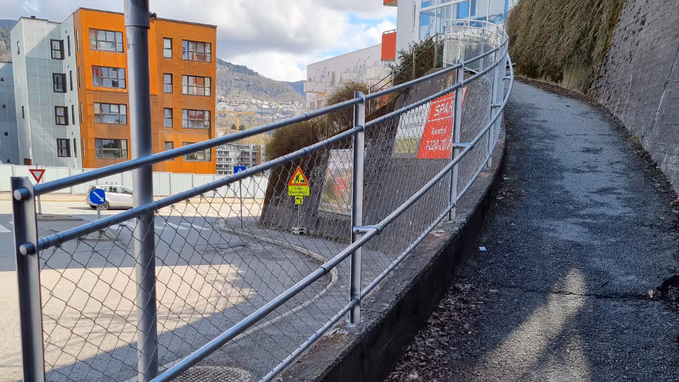 Rullebeskyttende rekkverk ved en asfaltert sti langs et stein- og gjerdeområde i et boligområde.