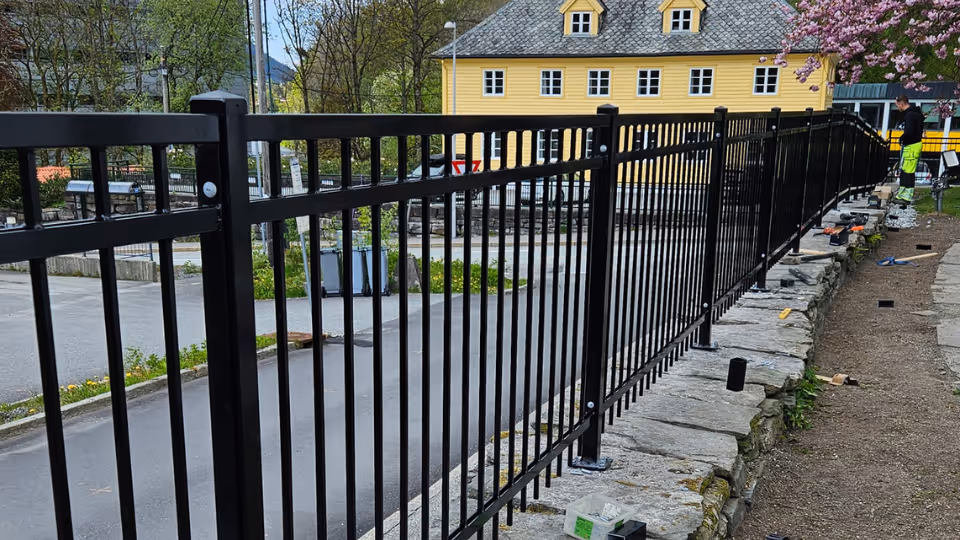 Sort smijernsrekkverk på lav mur i stein langs gate, med gult hus og blomstrende tre i bakgrunnen.