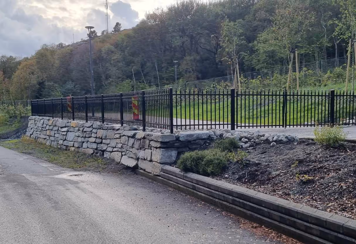 Sort metall rekkverk på en steinmur langs en vei med grønt gress og trær i bakgrunnen.