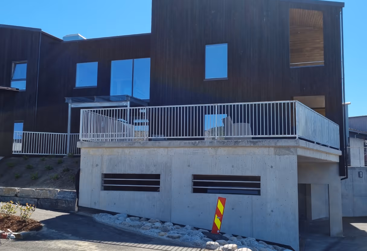 Moderne hus med mørk trefasade og betongkjeller med hvitt metallsikkerhetsrekkverk på terrassen.