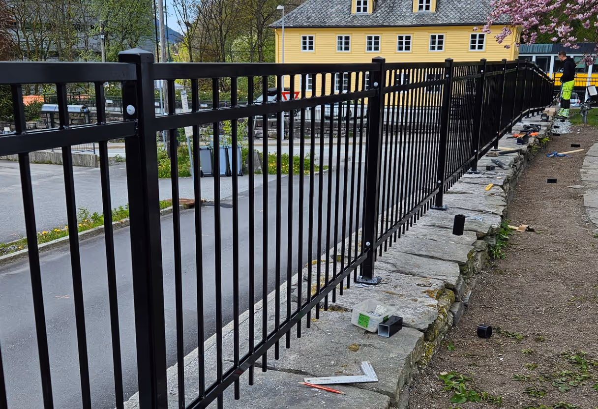 Montert svart metallrekkverk langs steinmur med arbeidstaker i bakgrunnen.