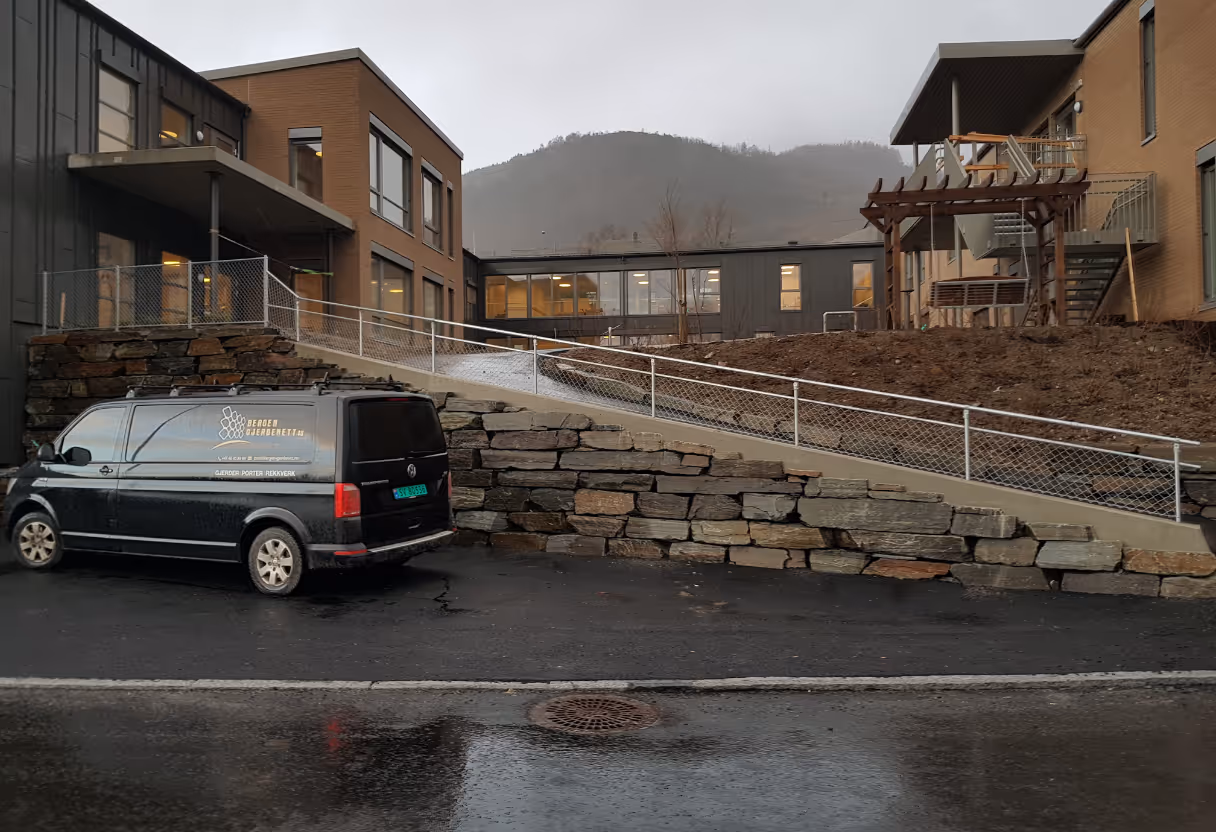 Rullestolrampe med hvitt rekkverk mellom to bygninger på vått vær, med svart varebil parkert foran.