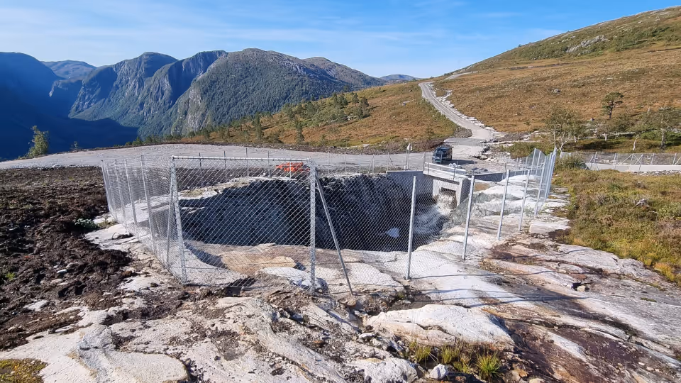 Sikringsgjerde rundt sikret grøft i fjellandskap med vei og fjell i bakgrunnen.