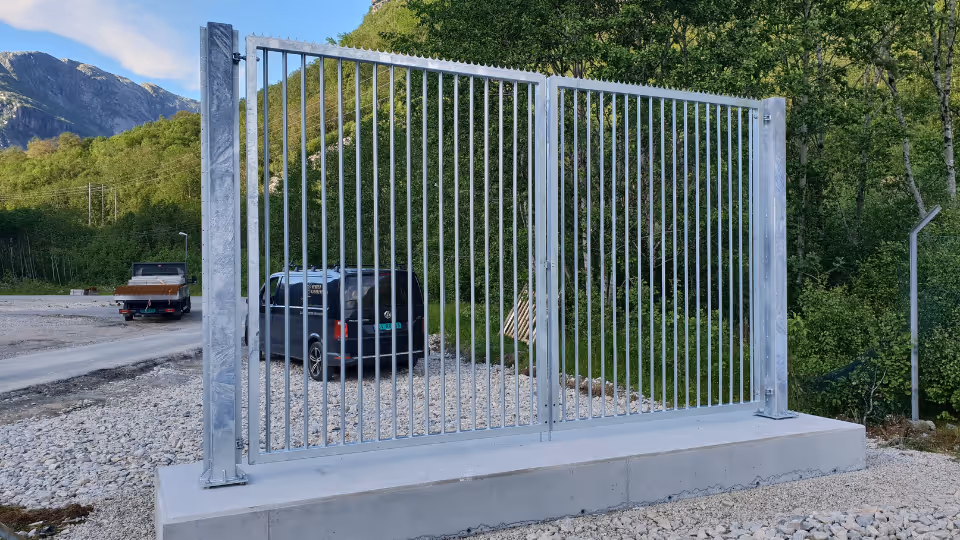 Metallport med vertikale stenger på betongfundament nær en skog og fjell.