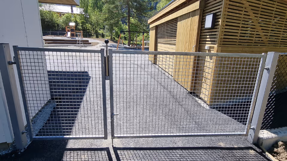 Metallgjerde med dør foran inngang til en barnehage med lekeplass i bakgrunnen.