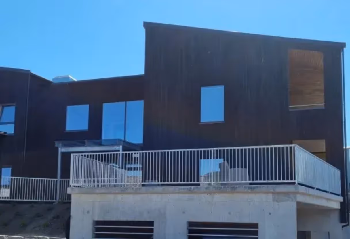 Moderne hus med stor balkong og hvitt rekkverk over betonggrunnmur under klar blå himmel.