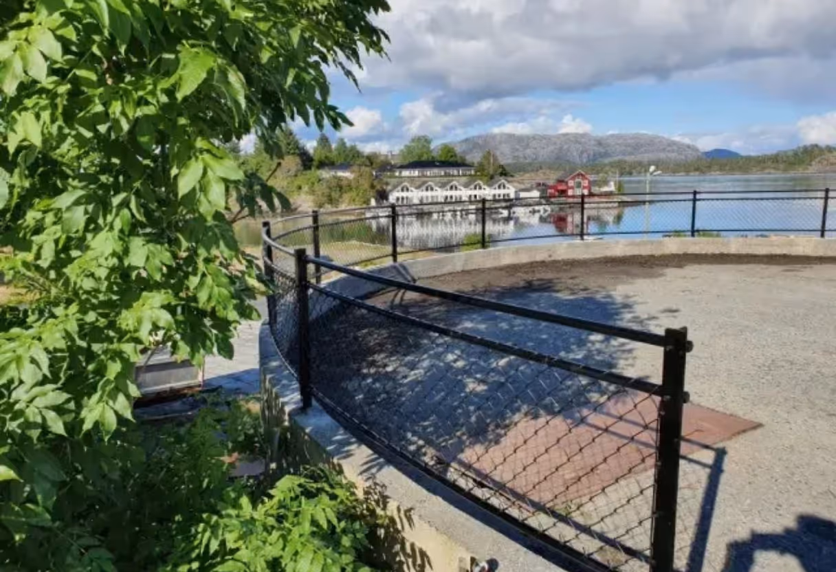 Svart metallrekkverk med netting rundt sirkulært område ved vann med trær og bygninger i bakgrunnen.