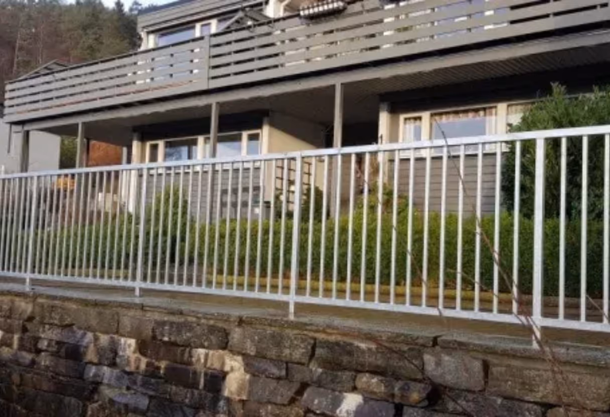 Hus med terrasse omsluttet av hvitt metallrekkverk og steinmur i forgrunnen.