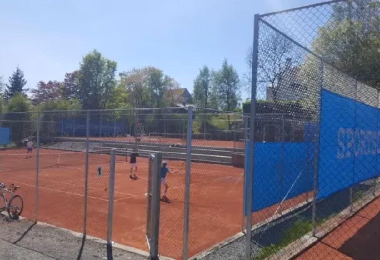 Tennisbane med to spillere i aktivitet bak gjerde under klar blå himmel.