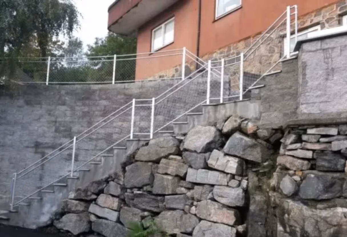 Betongtrapp med hvitt rekkverk og steinmur under, ledende til et bolighus.
