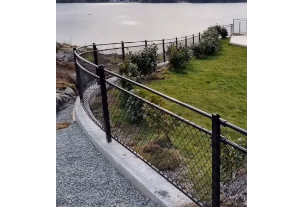 Sort rekkverk i metall med netting ved grønt område og vann i bakgrunnen.