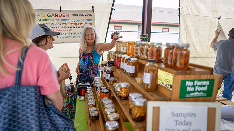 A friendly vendor offers homemade preserves and fresh produce at the Winnsboro Farmers Market. Shoppers browse colorful jars, local herbs, and natural foods that celebrate healthy living and the charm of East Texas.