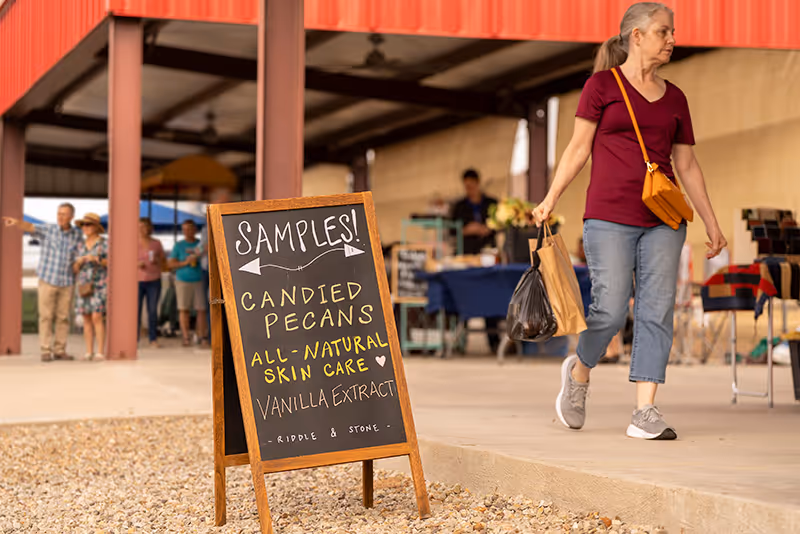 A welcoming scene at the Winnsboro Farmers Market featuring a display for samples of candied pecans, all-natural skincare, and vanilla extract from Riddle & Stone. Shop local and enjoy handmade East Texas goods.