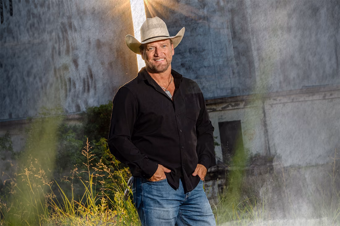 Man wearing a cowboy hat, black shirt, and jeans stands in a grassy area with a smile and hands in pockets.