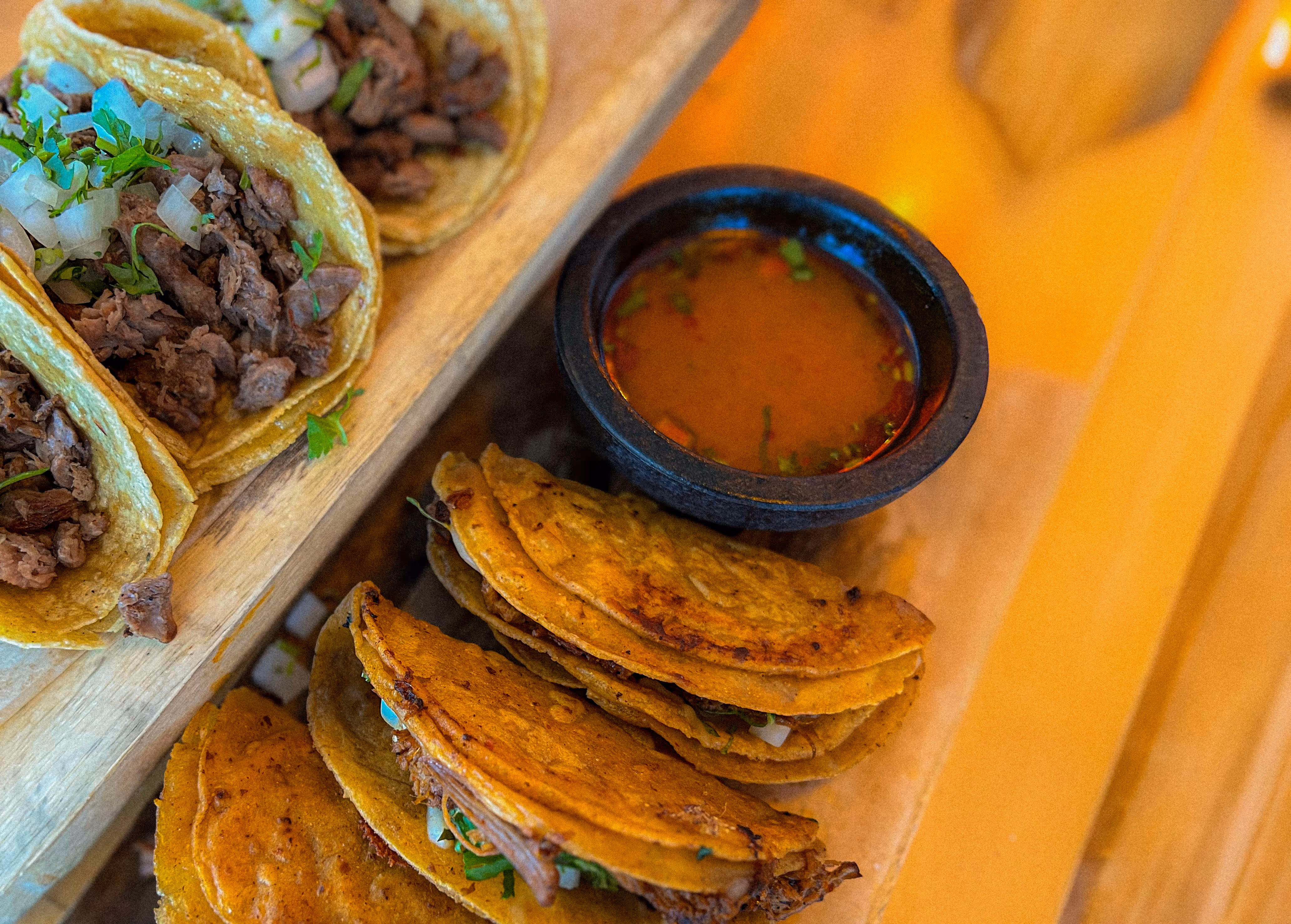 Close-up of authentic Mexican street tacos—traditional carne asada tacos and birria-style tacos—served on a wooden board with dipping consommé, available at Osteria del Mar.