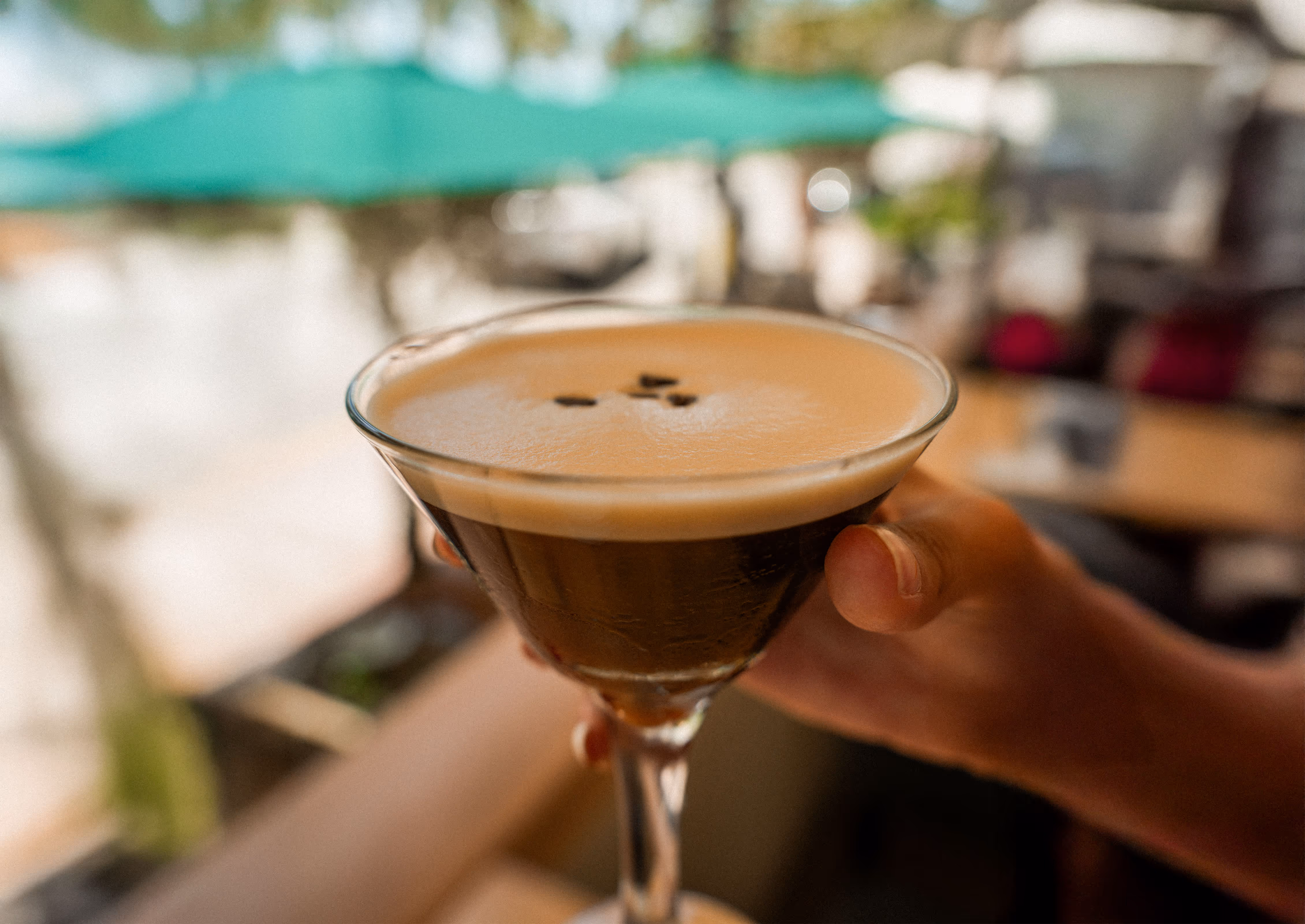 Hand holding an Espresso Martini cocktail garnished with coffee beans against a bright, sunny outdoor dining background at Osteria del Mar Miami Beach.