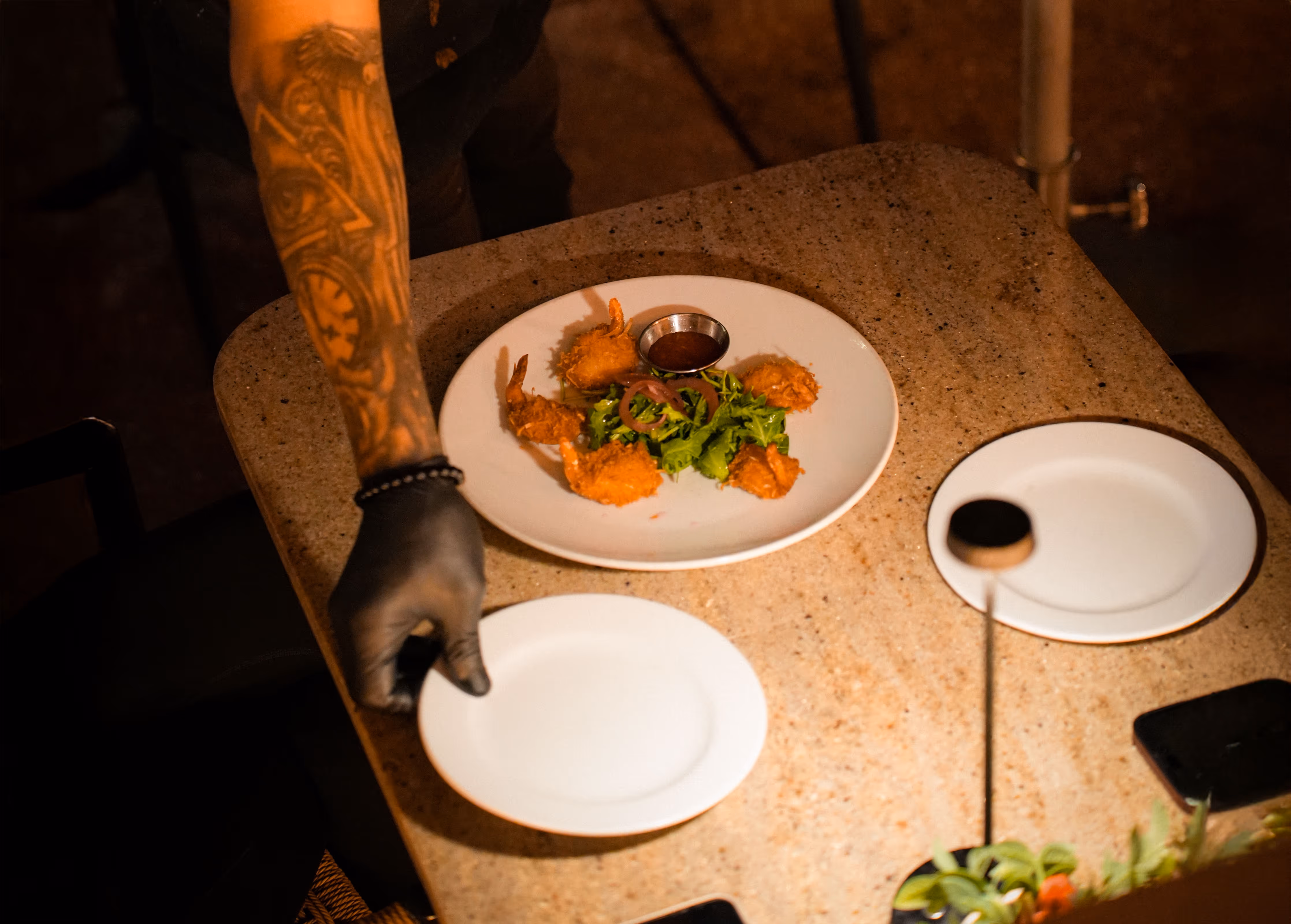 Close-up of a hand in a black glove serving a plate of fried shrimp appetizer with greens and a dipping sauce on a marble table at Osteria del Mar Miami Beach.