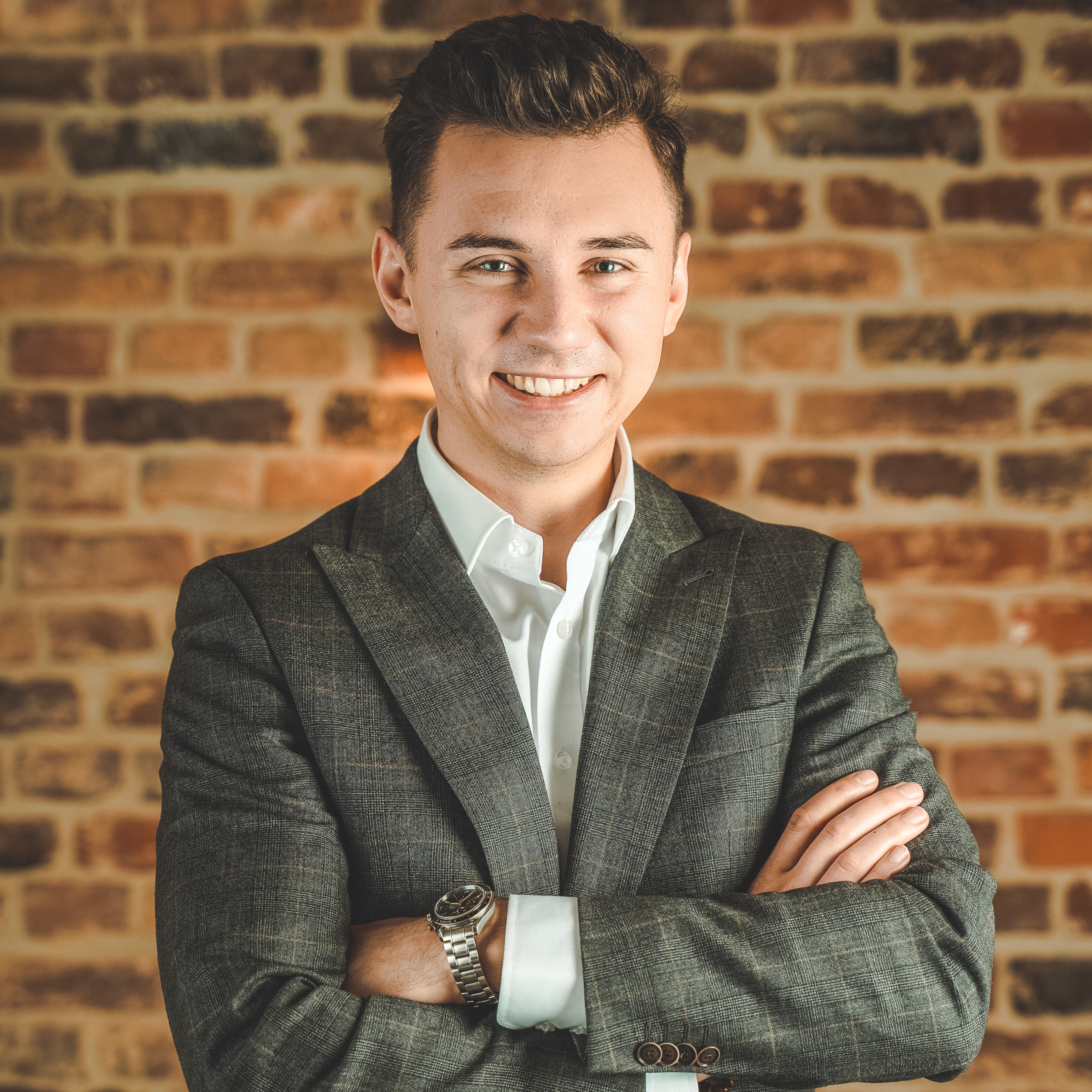 Young man in a gray plaid suit jacket and white shirt smiling with arms crossed in front of a brick wall.
