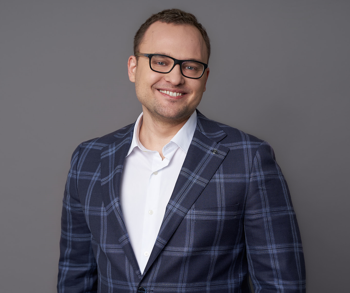 Smiling man wearing glasses and a blue plaid blazer over a white shirt against a gray background.