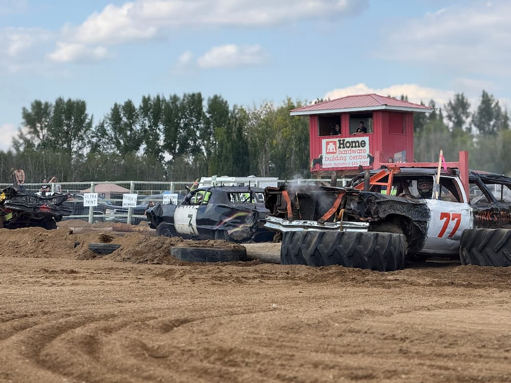 Two damaged demolition derby cars  on a dirt track near a judging stand.