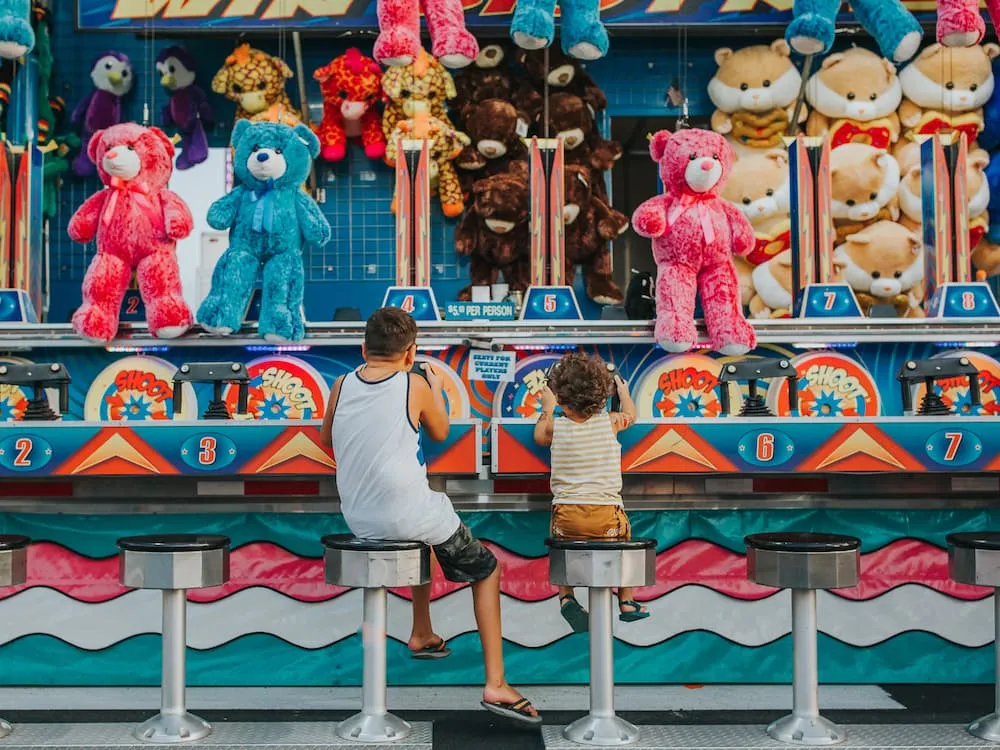 Two children sitting on stools playing a water gun shooting game at a carnival booth with large colorful stuffed animal prizes.