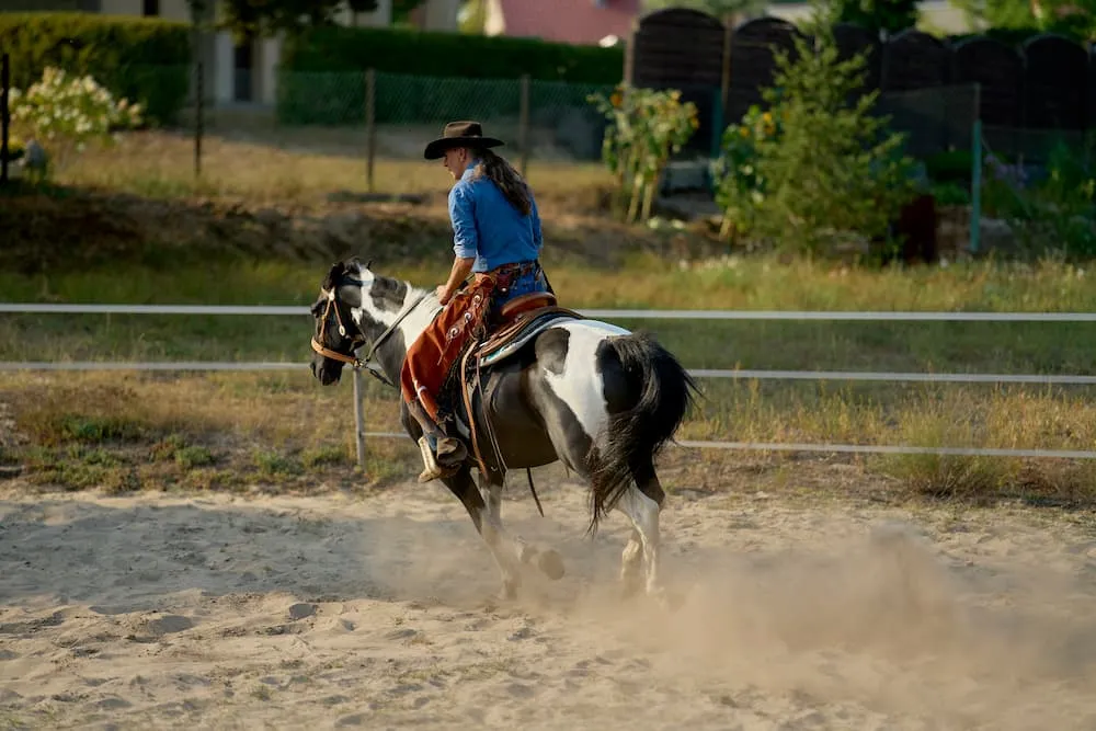 Woman in a blue shirt and cowboy hat riding a black and white horse on a sandy outdoor arena.