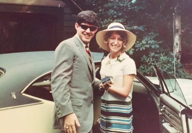 Smiling couple in vintage 1960s attire standing beside classic convertible car outdoors.
