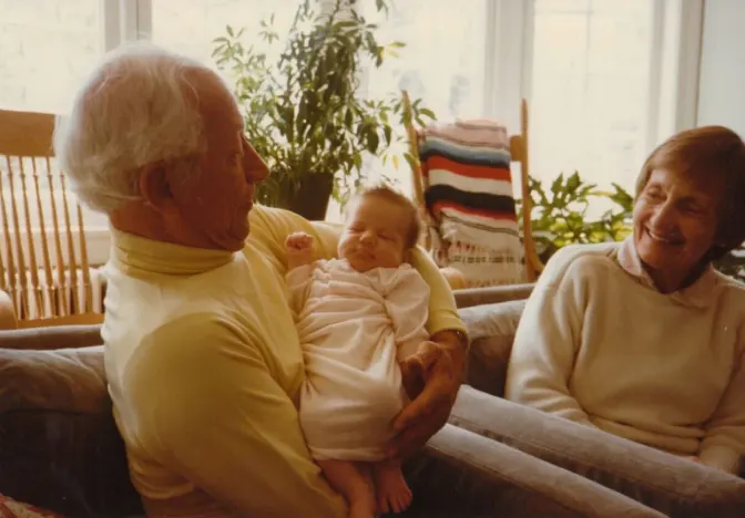 Older man holding infant while sitting with older woman on couch near sunny window and potted plants.