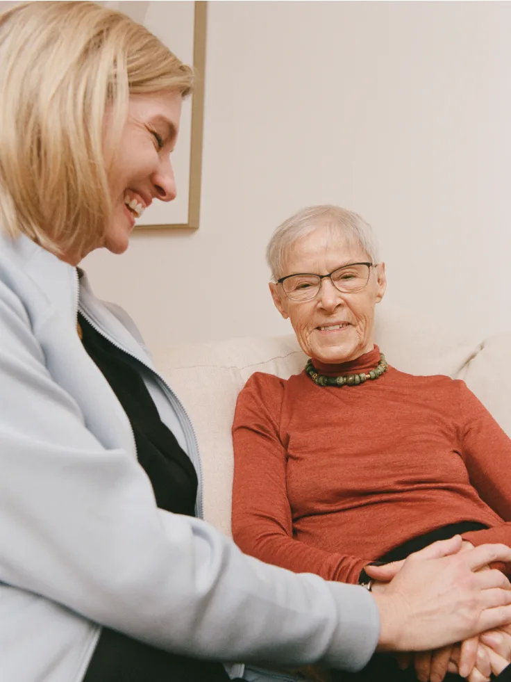 Smiling caregiver in white coat holding hands with senior woman in red shirt during supportive consultation.