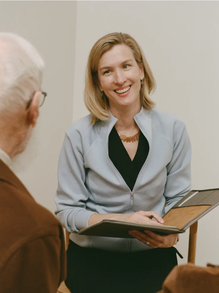 Smiling caregiver in light gray jacket speaking with elderly client while holding clipboard.