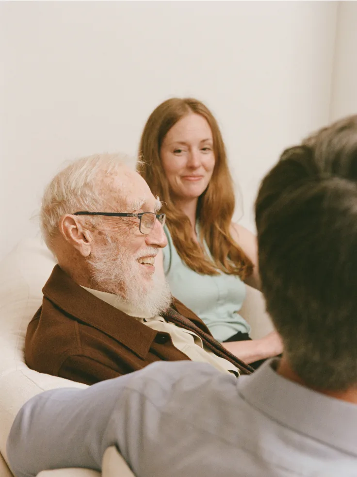 Senior man with glasses smiling during conversation with caregiver and another person in home setting.