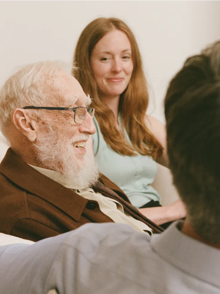 Smiling senior man with glasses and white beard beside caregiver in light blue shirt providing compassionate support.