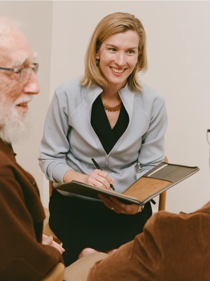 Care manager in light blazer taking notes while meeting with older clients in home setting.