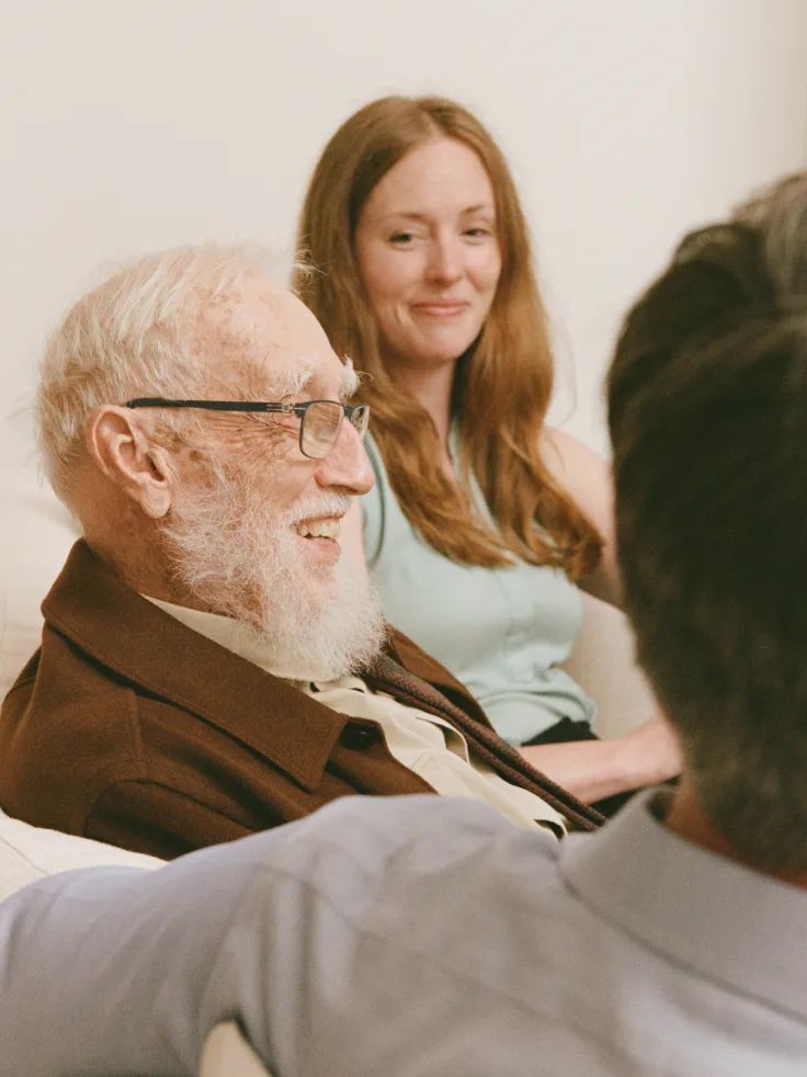 Senior man with glasses smiling during conversation with caregiver and family members.