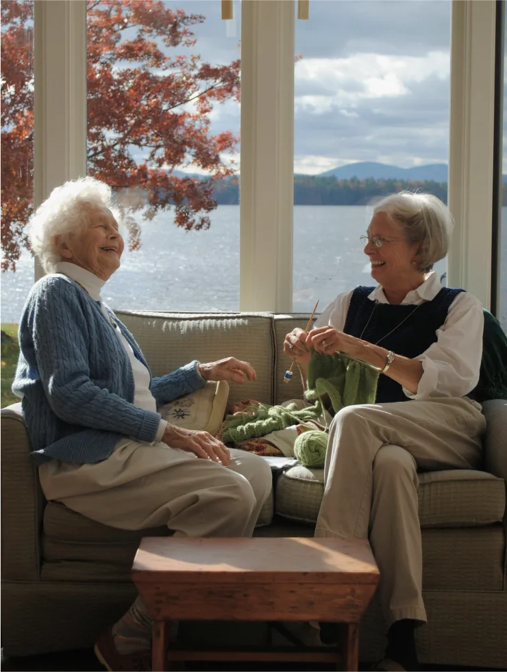 Two older women enjoy a meal together on a couch by a lake window with fall foliage and mountains visible outside.
