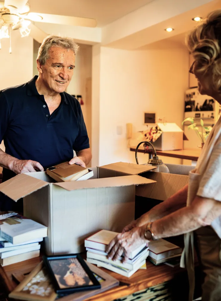 Senior man in navy polo smiling while packing boxes with older woman in warm, well-lit home interior.