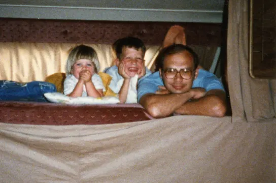 Older man with two young children smiling in bed together in a cozy home setting.