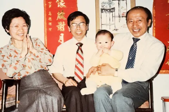 Four people posing together indoors with red decorative banners in background, including woman, two men, and young child.